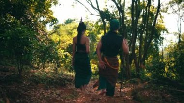 An Asian couple walking together in traditional green costumes inside the forest while the man holding a stick beside the woman during the morning