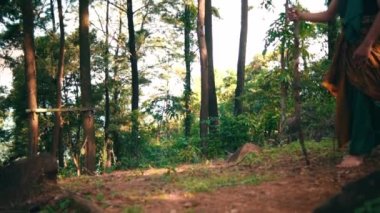 A Man with green clothes and a stick walking through the forest while visiting someone in the village during the morning