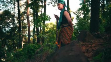 A Man with green clothes and a stick walking through the forest while visiting someone in the village during the morning