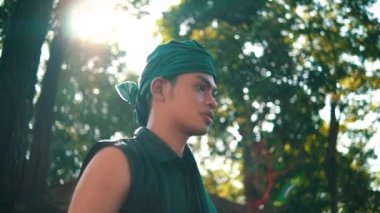 An Asian man in a green bandana and green clothes smiling while looking for someone in the forest during the daylight