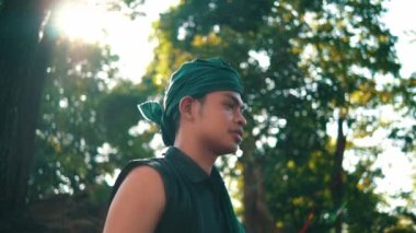 An Asian man in a green bandana and green clothes smiling while looking for someone in the forest during the daylight