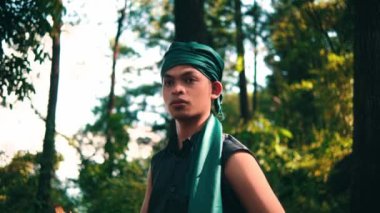 An Asian man in a green bandana and green clothes smiling while looking for someone in the forest during the daylight