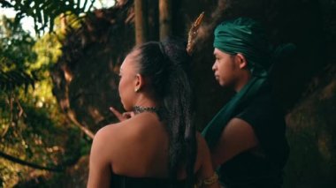 An Asian couple hangs out together in the forest while wearing a traditional green dress and talking to each other while enjoying the view during the daylight