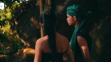 An Asian couple hangs out together in the forest while wearing a traditional green dress and talking to each other while enjoying the view during the daylight