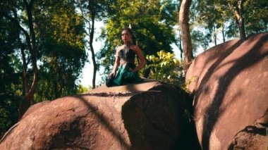 A beautiful Asian woman in a green costume sitting on a big rock while visiting the forest lot of trees inside the village