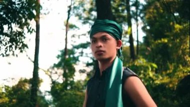 An Asian man in a green bandana and green clothes smiling while looking for someone in the forest during the daylight