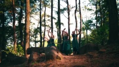 Bir grup Asyalı kadın, köydeki festivalde öğle vakti ormanın içindeki ağacın yanında yeşil elbiseler içinde dans ediyorlar.