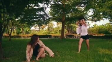 a group of women in shirts dancing with their friends in the middle of a park full of shady trees during the day
