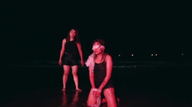 an Asian woman with her eyes and hands bound by a white cloth with a dirty body sitting on the wet beach sand at night