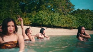a group of Asian women in brown clothes is enjoying the blue sea water by dancing with their friends on the beach on an island in the morning