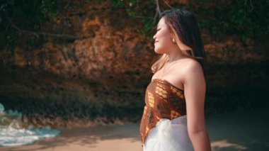 An Asian woman with blonde hair walks towards the beach with beautiful white sand with a smile while on vacation on an island during the day