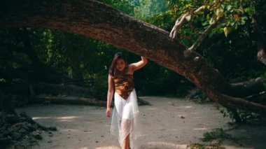 An Asian woman in a brown shirt walks past a large tree trunk on the beach sand while on vacation on an island that has lots of green trees during the day