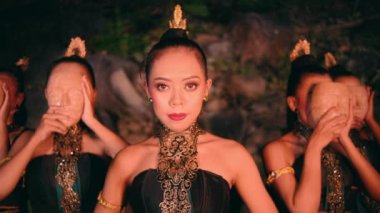 a group of Asian women in green traditional clothes wearing bamboo masks together while standing among the rocks at night