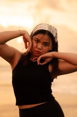 an Asian teenager wearing a white bandana and black shirt with a sexy expression on the beach sand in the afternoon