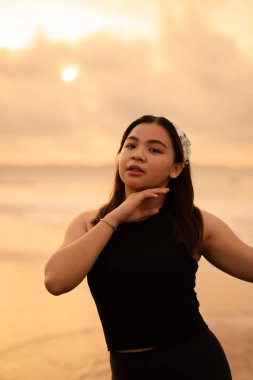 a glamorous Asian woman in black clothes and a white bandana standing in front of the waves on the beach in the morning