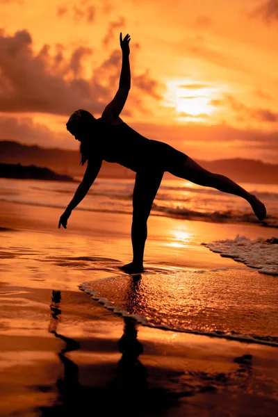 silhouette of an Asian woman dancing ballet with great flexibility and a view of the waves behind her at sunset