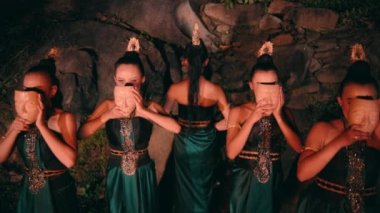 an Asian woman walking among a group of women wearing bamboo masks with smiling faces in front of the rocks at night