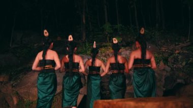 a group of Asian women standing together in front of big rocks while holding bamboo masks in their hands with flat expressions in the middle of the forest at night