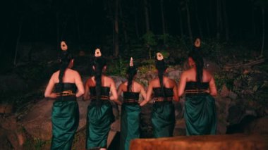 Bir grup Asyalı kadın, gecenin bir yarısı ormanın ortasında ellerinde bambu maskeler ve düz ifadelerle büyük kayaların önünde bir arada duruyorlar.