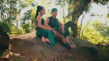 Asian woman and man are sitting on a rock wearing green dresses enjoying the view during the day