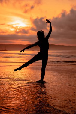 silhouette of a very slender ballerina doing ballet practice alone on the seashore with waves crashing at her feet at sunset