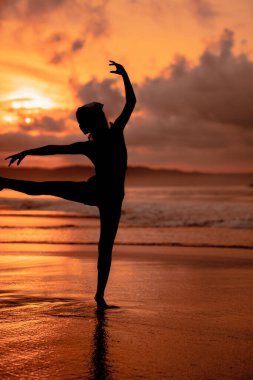 silhouette of a very slender ballerina doing ballet practice alone on the seashore with waves crashing at her feet at sunset