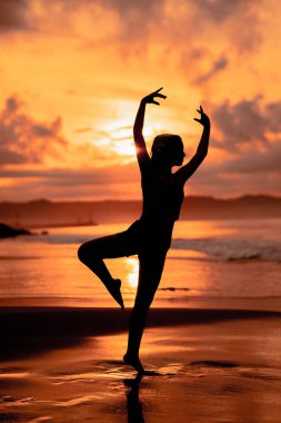 A ballerina with a silhouette shape performs ballet movements very flexibly on the beach with the waves crashing in the afternoon
