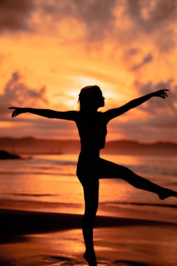 A ballerina with a silhouette shape performs ballet movements very flexibly on the beach with the waves crashing in the afternoon