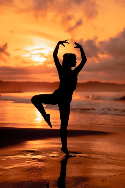 A ballerina with a silhouette shape performs ballet movements very flexibly on the beach with the waves crashing in the afternoon