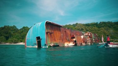 a shipwreck stranded in front of an island full of green trees and blue sea during the day