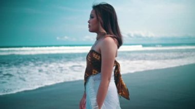 An Asian woman in a white dress walking alone on the beach with a sad expression and the sea waves behind her during the day