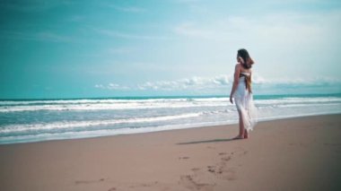 An Asian woman in a white dress is standing on the beach while looking at the beautiful view of the blue sea and the waves during the day