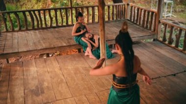 a group of Asian female dancers in green costumes dance together in front of a sad woman in a bamboo house around the forest