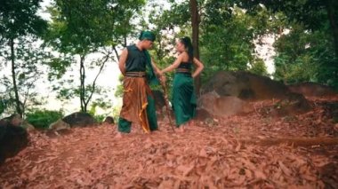 An Indonesian man and woman dancing together wearing green clothes on a ground full of green leaves in the middle of a forest in the morning