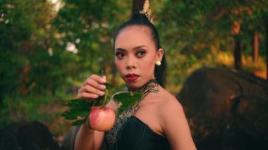 an Asian woman holding her fruit with an angry expression while wearing a green dress in the middle of the forest in the morning