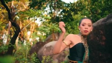 An Asian woman approaches someone with an angry expression while wearing green clothes and makeup in the middle of the forest in the morning
