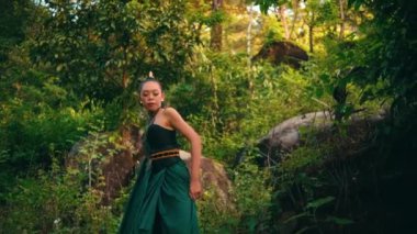 An Asian woman approaches someone with an angry expression while wearing green clothes and makeup in the middle of the forest in the morning