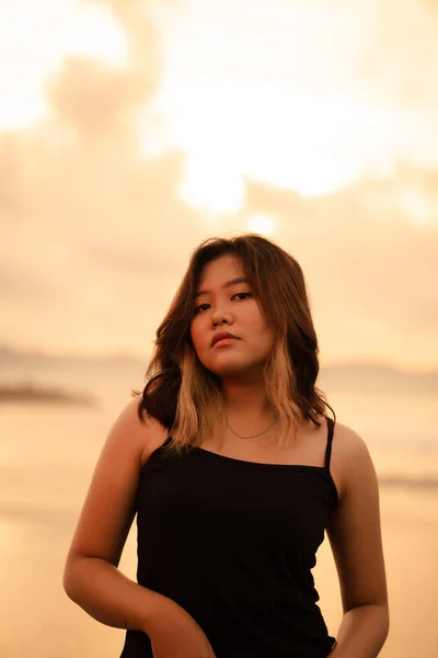 An Asian woman poses with a dirty and angry expression when wearing a black dress on the beach in the morning
