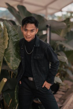 an asian boy in a black denim jacket posing like a naughty boy with a chain around his neck in a park in the afternoon