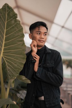 an asian boy in a black denim jacket posing like a naughty boy with a chain around his neck in a park in the afternoon