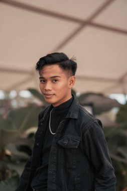 an asian boy in a black denim jacket posing like a naughty boy with a chain around his neck in a park in the afternoon