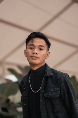 an asian boy in a black denim jacket posing like a naughty boy with a chain around his neck in a park in the afternoon