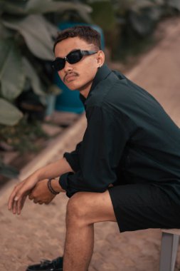 an Asian man with a mustache face wearing a shirt and sunglasses while hanging out with his friends in a cafe during the day