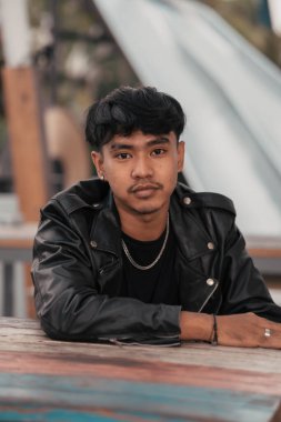an Asian man with a naughty face wearing a chain necklace and a black leather jacket in a cafe during the day