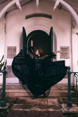 An elegant witch flicks her all black dress in the air in front of the cemetery gates after the Halloween festival begins in the afternoon
