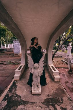 a young witch in a black dress is standing between the tombstones shaped like an angel while visiting the cemetery in the afternoon