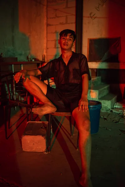 a gay man wearing black and posing with a chair in an old house with a red light at night