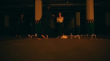 a woman with blonde hair and black clothes is reciting a ritual incantation surrounded by men running around the pentagram altar at night