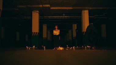 a woman with blonde hair and black clothes is reciting a ritual incantation surrounded by men running around the pentagram altar at night