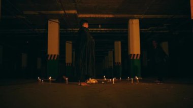 a woman with blonde hair and black clothes is reciting a ritual incantation surrounded by men running around the pentagram altar at night